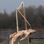 Fotografia kolorowa przedstawia rzeźbę wykonaną z drewna lipowego. Rzeźba jest organiczną formą zdeformowanego ciała wspieraną przez geometryczną konstrukcję drewnianą. W tle rzeka i drzewa.