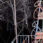 Fotografia ukazująca fragment instalacji „Ogrody starości” oraz fragment instalacji „Olbrzym”. Stare zabawki z placu zabaw na tle wybielonych drzew jabłoni ustawionych w metalowych piramidach.