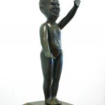 Rzeźba z brązu przedstawiająca figurę lalki-zabawki z realistycznym portretem i uniesioną lewą ręką.